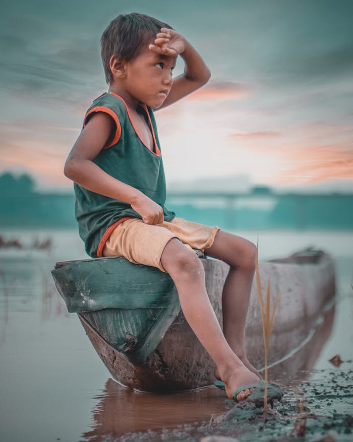 about-us A young boy sits on a wooden boat by a misty riverside in Nepal, gazing into the distance.